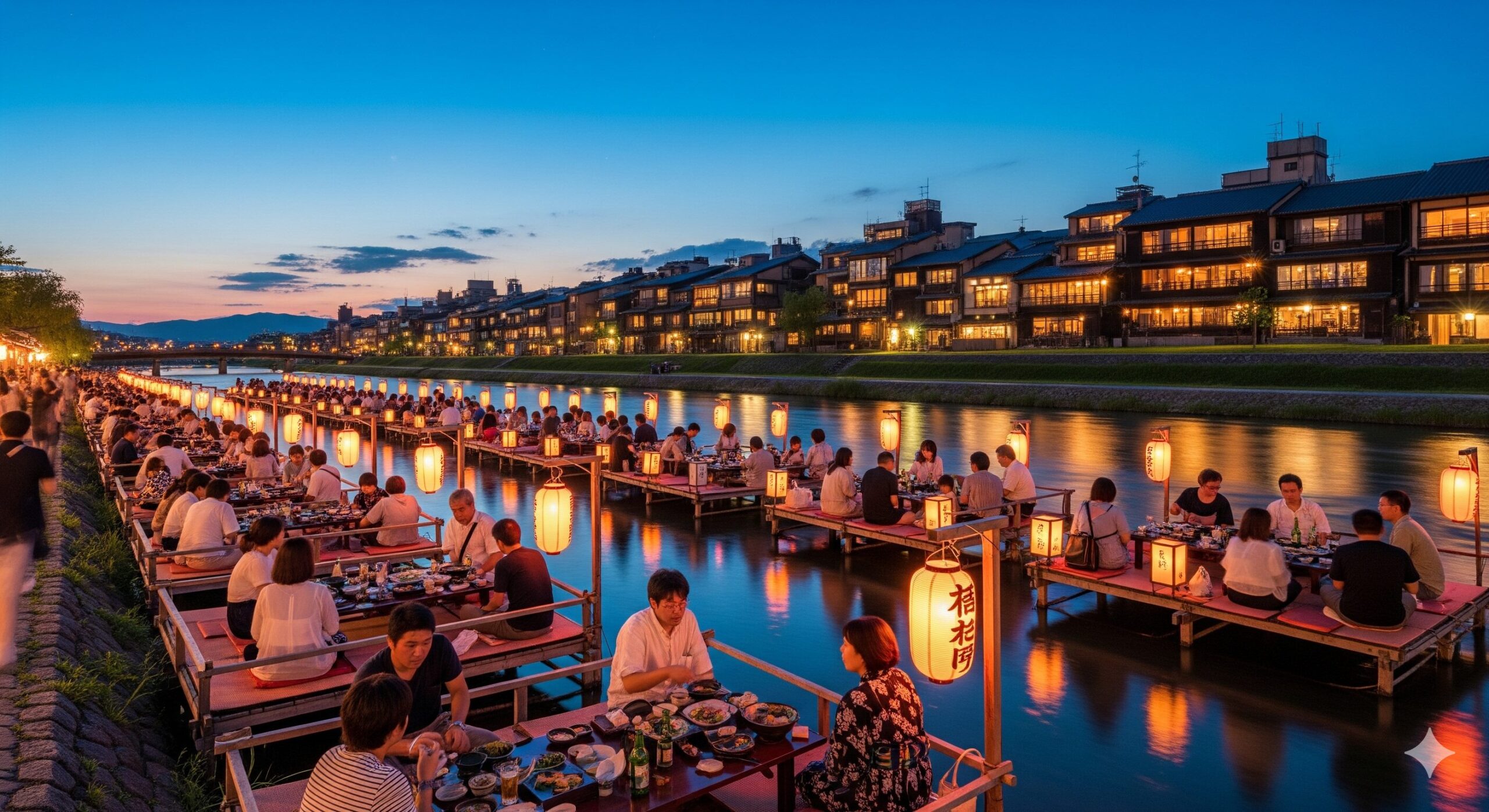 京都の鴨川沿いに設置された夏の納涼床の風景。多くの人々が川床で食事や談笑を楽しんでおり、川面には夕日が反射している。対岸には京都の街並みが広がる。