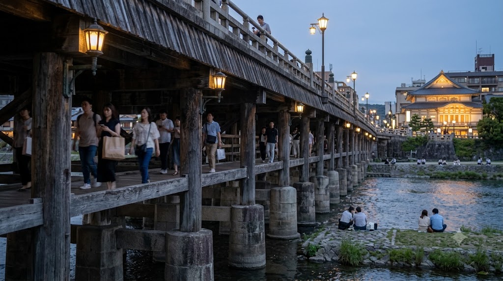 京都の三条大橋。木造の古風な橋を多くの人が行き交い、その下の鴨川右岸（西側）の河川敷には、等間隔に座る人々が見える。夕暮れ時。