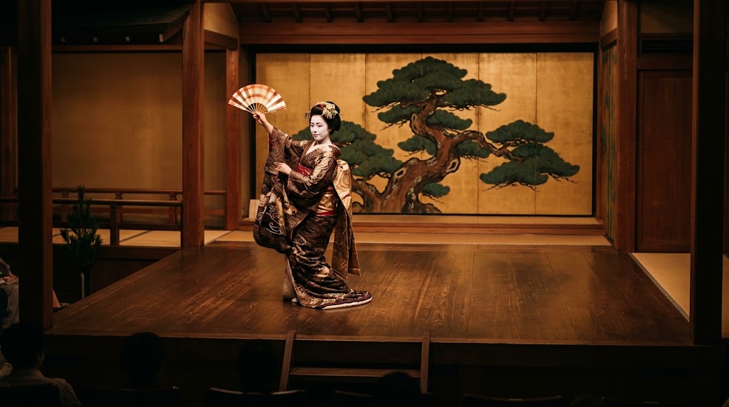 上七軒歌舞練場の歴史ある木造舞台で、伝統的な日本舞踊を披露する格式高い芸妓。豪華な意匠の着物を身にまとい、伝統的な日本髪に華やかな髪飾り(簪)をあしらい、手にした扇子(舞扇)で優雅な所作を見せる舞台パフォーマンス。背景には大きな松が描かれた金色の障壁画があり、劇場内の温かみのあるスポットライトに照らされている。