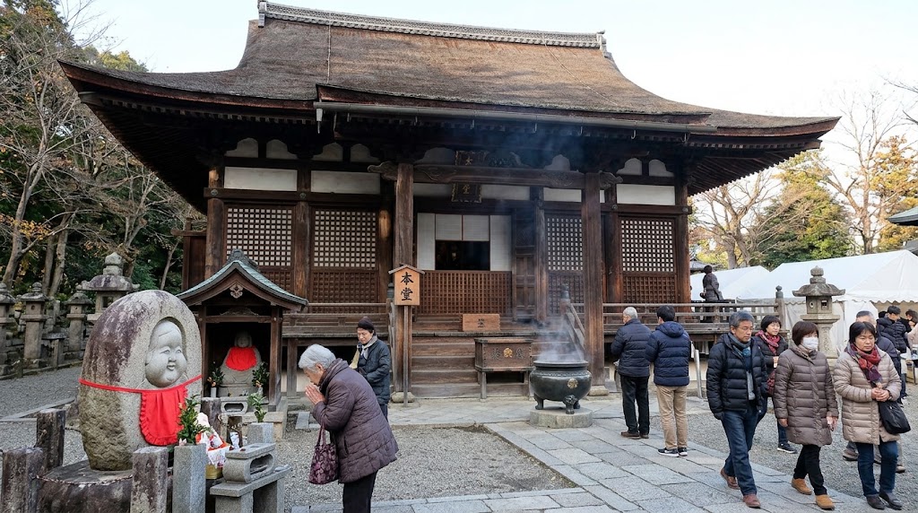 鎌倉時代（1227年建立）の面影を残す千本釈迦堂（大報恩寺）の国宝本堂。手前左側には、伝説の「おかめ」のふくよかな顔が彫られた石碑（おかめ塚）があり、冬の装いをした参拝客が静かに手を合わせている。本堂の前では香炉から煙が立ち上り、多くの参拝客が境内を行き交う様子。