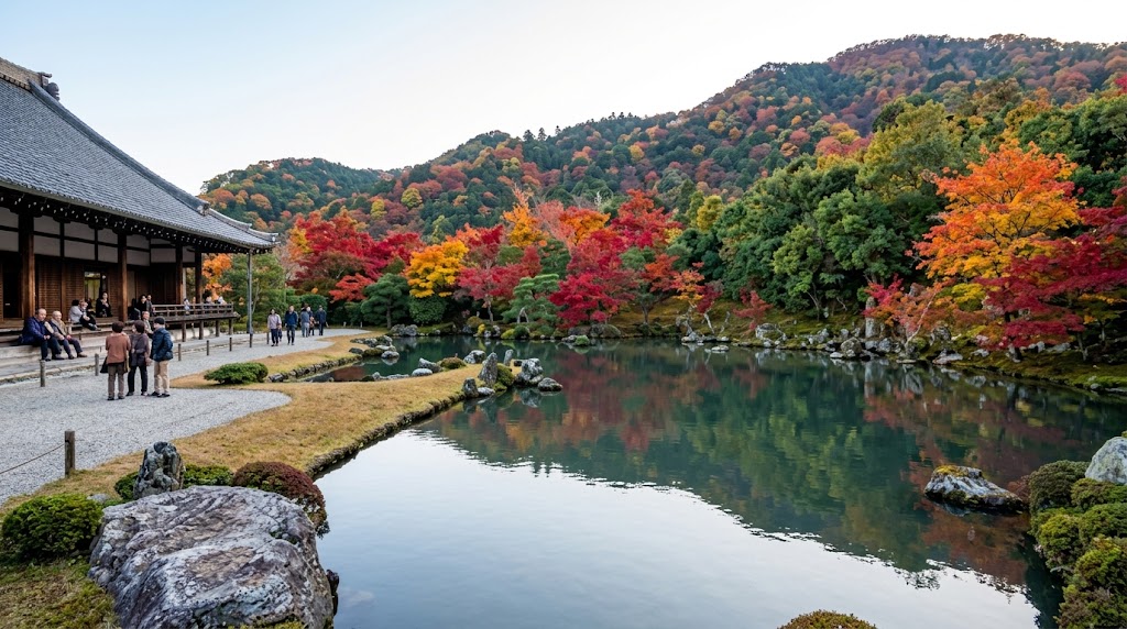 京都・嵐山にある天龍寺の曹源池庭園。赤や黄色の紅葉、池、そして背後に広がる色づいた嵐山の山並みを、お堂の縁側や庭園から眺める人々。